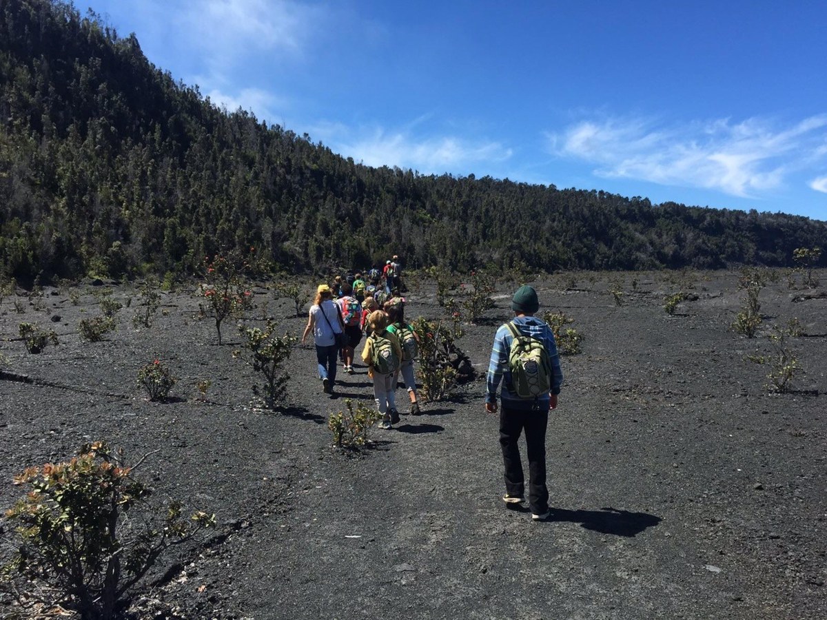 孩童與健行者走在基拉韋亞火山口底部的黑色熔岩地面上，遠處是火山口邊緣的森林