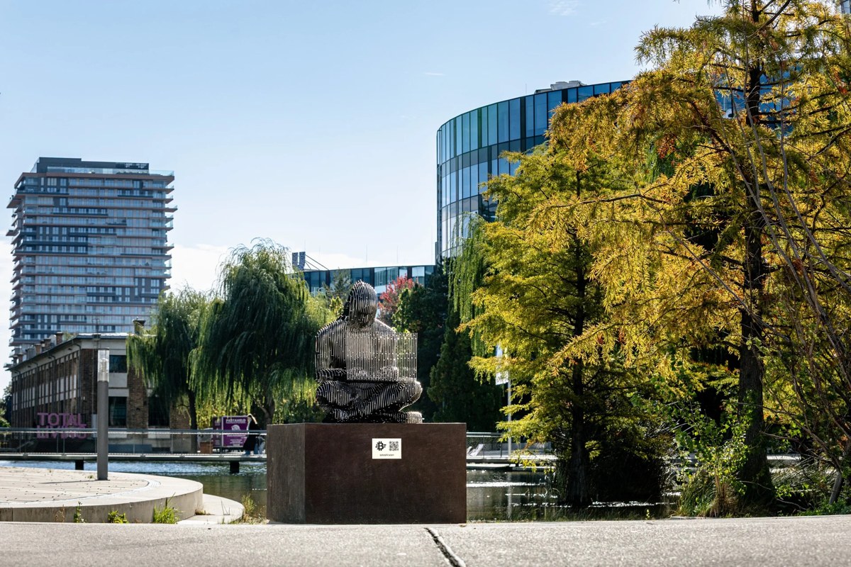維也納 Viertel Zwei 湖畔的中本聰不鏽鋼雕像，坐在鏽紅色鋼製底座上，周圍是秋天的樹木和辦公大樓