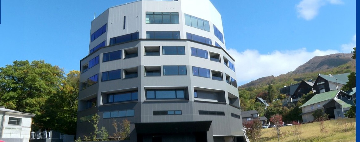 藍天下一棟多角形的灰白色建築,背後是山和樹林