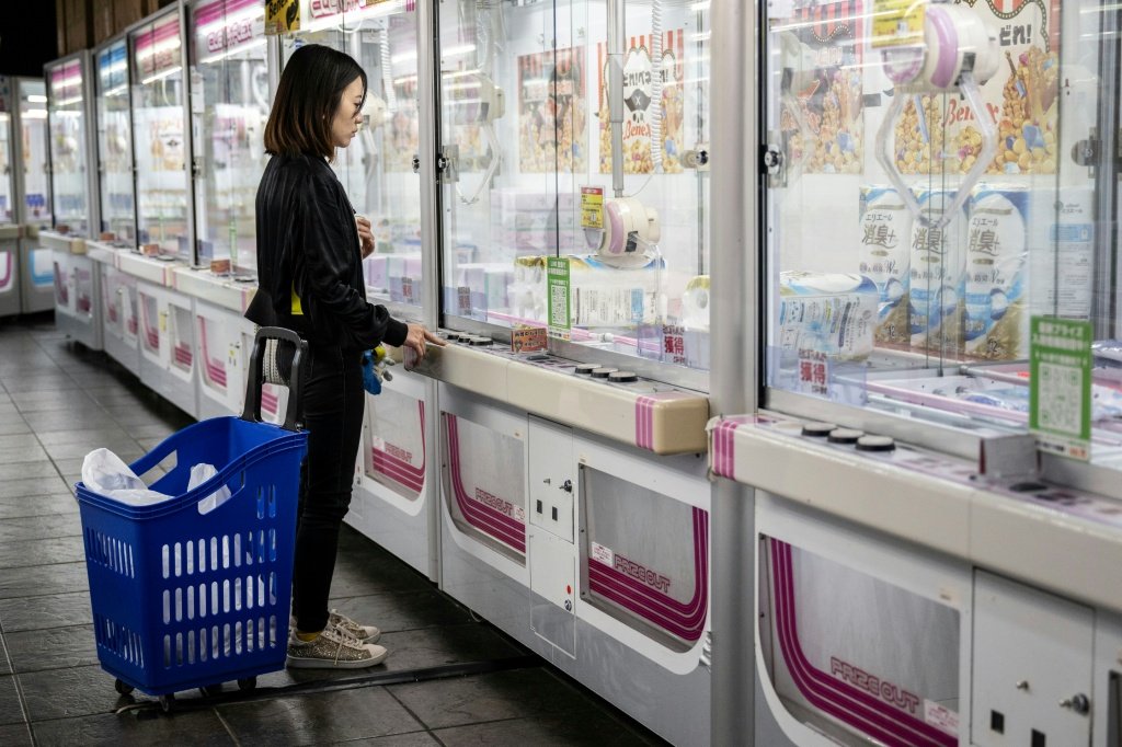 川崎遊樂場內，一位推著藍色購物籃的女子站在裝滿日用品的夾娃娃機前